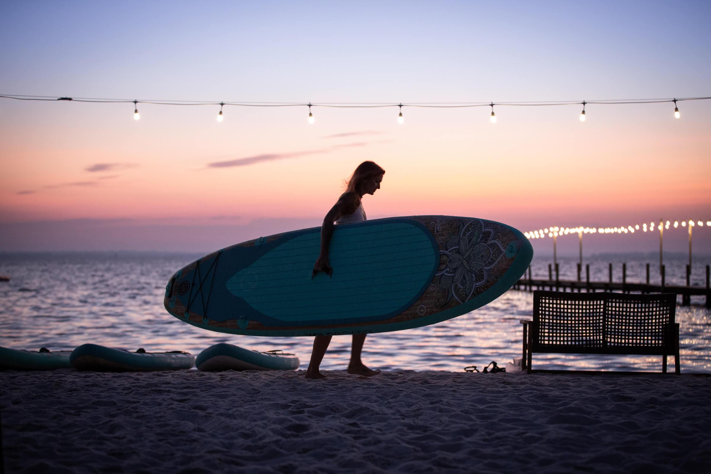 Yolo Board | Woman Carrying Sup On Beach Along Ocean Shore At Dusk